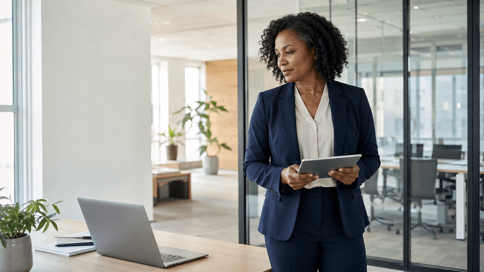 A finance executive holding a tablet in a bright modern office, calm and composed, a laptop on the desk.